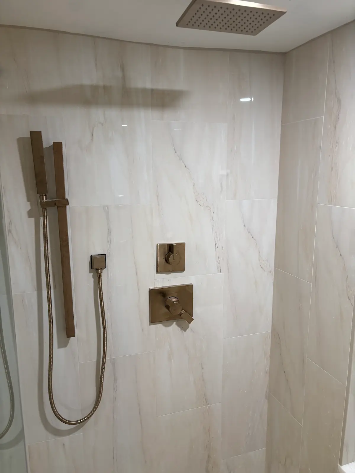 Modern brushed bronze shower hardware including a square rain showerhead, a vertical handheld spray wand, and sleek square control valves on marble tile walls.