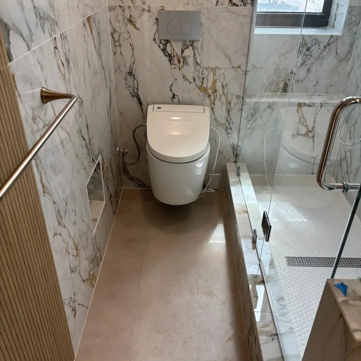 Modern luxurious bathroom interior with large-format white and gold marble tile walls, a wall-mounted floating toilet, and a glass shower door revealing a tile floor with a linear drain.