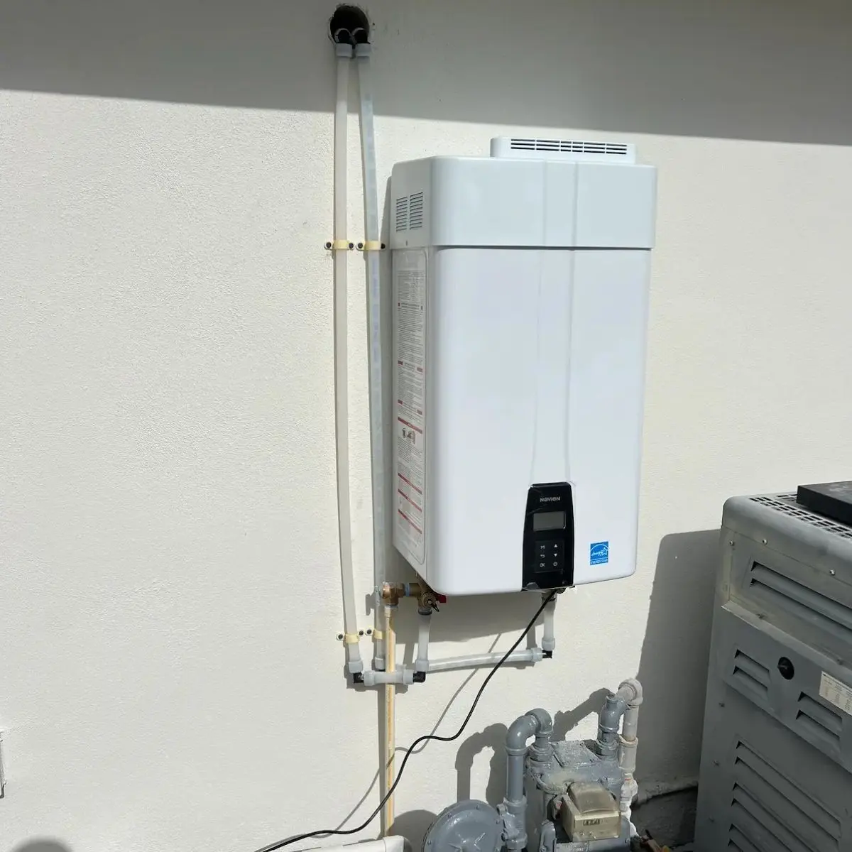 A white Navien tankless water heater unit mounted externally on a textured white stucco house wall above a gas meter setup.