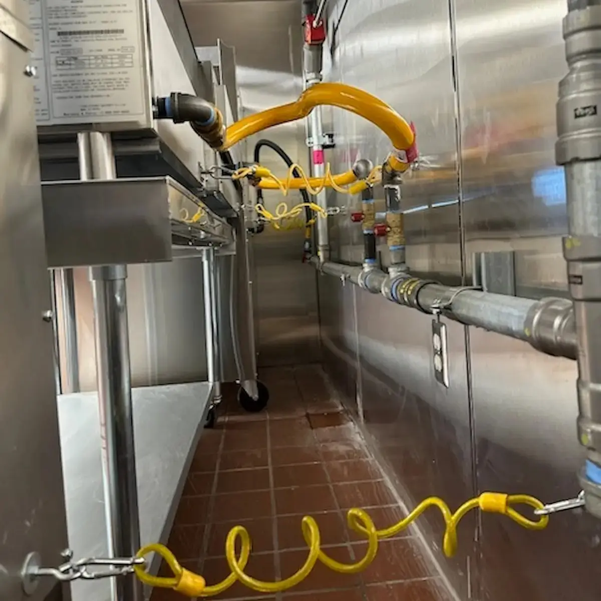 Stainless steel commercial kitchen line showing gas pipes, plumbing utilities, and flexible yellow lines running behind heavy-duty restaurant equipment.