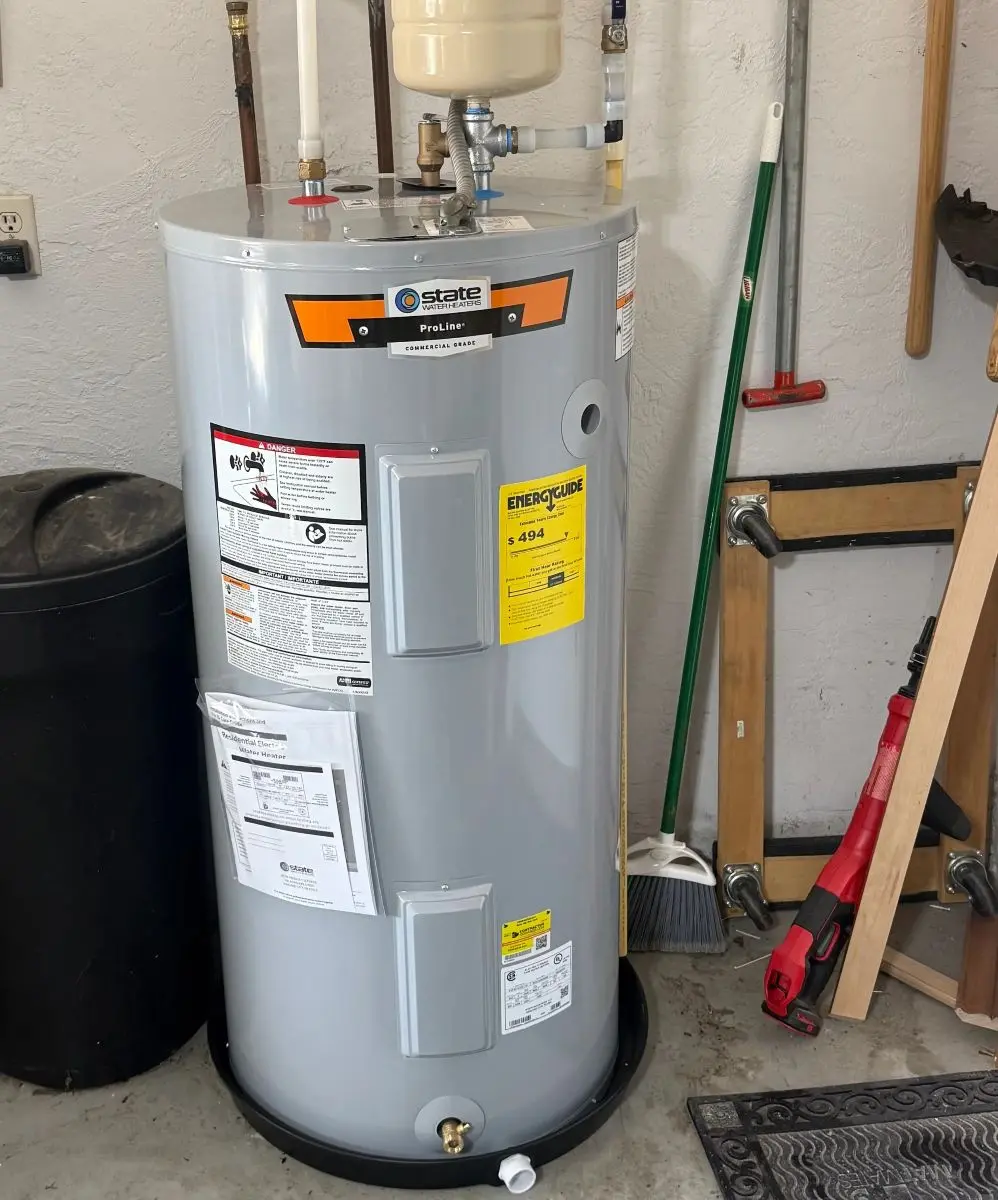 A tall silver State ProLine water heater tank standing on a garage floor in a black plastic overflow pan, featuring clean pipe tie-ins and a small beige expansion tank on top.